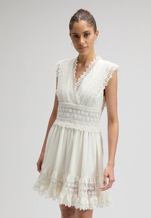 Donna con capelli raccolti in uno chignon che indossa un abito bianco senza maniche in pizzo con scollo a V e orlo arricciato, in piedi davanti a uno sfondo chiaro e uniforme.