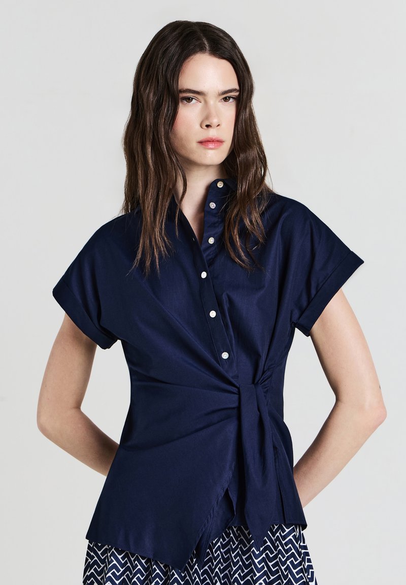 Donna con lunghi capelli scuri che indossa una blusa blu navy a maniche corte con bottoni e cintura in vita, abbinata a una gonna a fantasia, mentre posa su uno sfondo neutro.