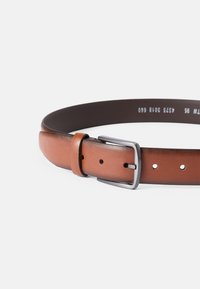 Καφέ δερμάτινη ζώνη με λεία υφή, διαθέτει ασημένιο μεταλλικό κούμπωμα και τρύπες για ρύθμιση. Η ζώνη έχει αντίθεση με σκούρο περίγραμμα.