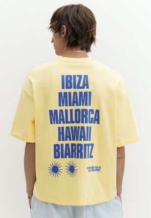 Osoba stojąca tyłem, ubrana w żółtą koszulkę z niebieskim napisem wymieniającym Ibiza, Miami, Majorka, Hawaje i Biarritz z grafikami słońca poniżej.