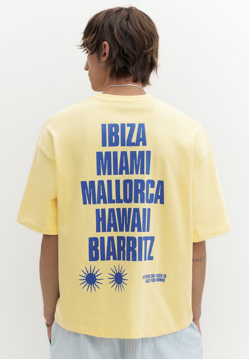Personne de dos portant un t-shirt jaune avec un texte bleu listant Ibiza, Miami, Majorque, Hawaï et Biarritz avec des graphismes de soleil en dessous.