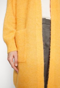 Gros plan sur une personne portant un cardigan jaune doux et tricoté sur un haut blanc et un pantalon foncé, avec une main près d'une poche avant.