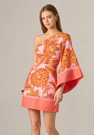 Kvinde med langt brunt hår iført en lyserød kjole med ét skulderstykke med store orange blomsterprint og en bred ærme på den ene arm.