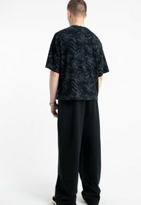 Osoba stojąca tyłem, ubrana w czarną, oversize'ową koszulę w szare fale oraz luźne czarne spodnie na białym tle.