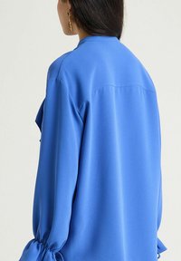 Blusa blu in tessuto liscio, con maniche lunghe e polsini arricciati, vestibilità comoda e design con giogaia posteriore.
