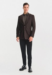 Mężczyzna stoi, nosząc ciemno-brązową marynarkę, wzorzystą koszulę, czarne spodnie i czarne, wypolerowane eleganckie buty na tle jednolitego białego tła.