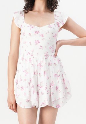 Donna che indossa un abito bianco a fiori rosa, scollatura quadrata, maniche arricciate e gonna fluente, in piedi con una mano sul fianco.