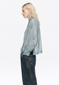 Jasnoniebieska, przezroczysta koszula na guziki z długimi rękawami, charakteryzująca się teksturowanym wzorem i luźnym krojem. Noszona z ciemnoniebieskimi dżinsami.