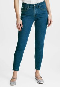 Úzké džíny v tmavě modrém denimu, s vysokým pasem, klasickým pětikapsovým designem a nenápadnými detaily v prošívání. Lemy ohrnuté kolem kotníků.