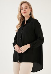 Fekete, hosszú ujjú gombos ing, amely puha textúrával, egyenes gallérral és magas-alacsony szegéllyel rendelkezik, finom fényű.