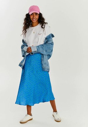 Kobieta w różowej czapce, białej bluzie, dżinsowej kurtce, niebieskiej spódnicy w wzory i białych tenisówkach, stojąca na jednolitym tle.