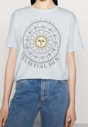 Osoba w jasnoniebieskiej koszulce z żółtą twarzą słońca, otoczoną kwiatami, gwiazdami oraz napisem „CELESTIAL SUN”, włożoną w niebieskie dżinsy.