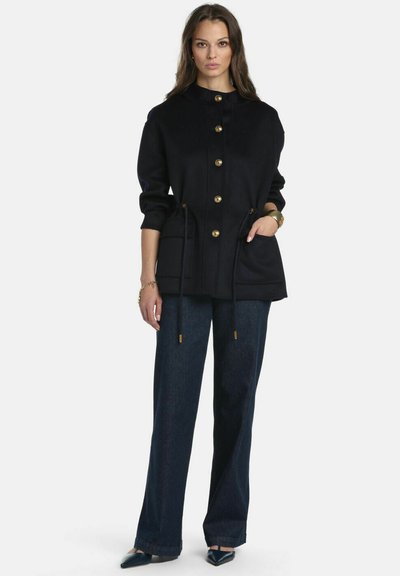 Giacca navy con bottoni dorati, vita regolabile con coulisse e tasche laterali. Abbinata a pantaloni di denim scuro e scarpe a punta.