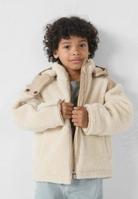 Chaqueta de forro polar beige esponjosa con un acento en los hombros de color marrón claro y detalles de cremallera. La modelo la lleva sin cerrar sobre una camiseta gris y vaqueros azul claro.