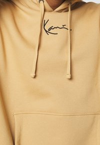 Sudadera beige con bolsillo frontal y cordones, que presenta un pequeño logotipo negro bordado en el centro del pecho.