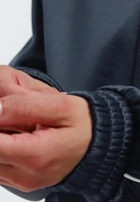 Dark blue zip-up jacket with ribbed cuffs; hands are clasped, showing smooth skin; no notable patterns or hardware visible.