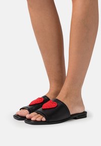 Black leather slide sandals featuring two red, textured heart accents on the upper with an open-toe design and flat sole.