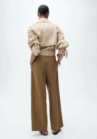 Femme debout les mains dans les poches, portant une veste beige courte avec des manches retroussées et un pantalon brun large, de dos sur fond uni.