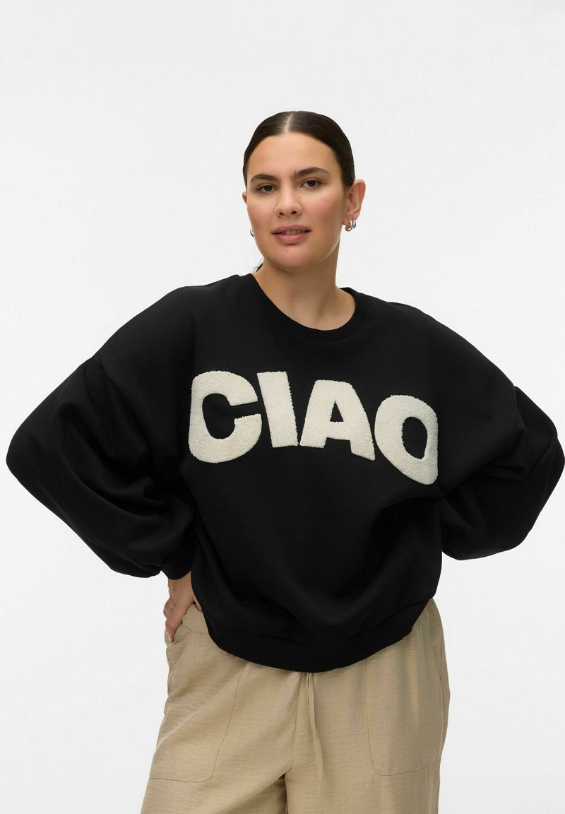 Femme aux cheveux foncés portant un pull noir surdimensionné avec le mot « CIAO » en grandes lettres blanches, un pantalon beige, posant les mains sur les hanches devant un fond blanc.