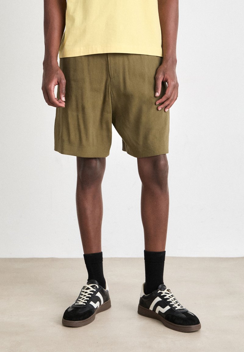 Olive green shorts made of lightweight fabric, with a relaxed fit. Paired with black sneakers featuring beige and white accents, and ribbed black socks.