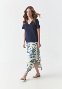 Top de pico en color marino combinado con una falda larga crema con patrones de hojas verdes y azules; sandalias que dejan los pies al descubierto, tejido casual y un diseño fluido.