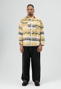 Geel en grijs patroonjack met geometrische vormen, gecombineerd met zwarte broek met brede pijpen en stevige zwarte schoenen. Soepele textuur.