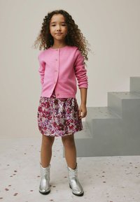 Rosa Cardigan mit goldenen Knöpfen, kombiniert mit einem glitzernden Rock mit floralem Muster in Pink und Lila. Silberne Cowboystiefel runden das Outfit ab.