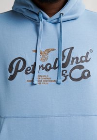 Světle modrá mikina s kapucí a šňůrkami, s výrazným černým a zlatým vyšívaným textem a logem orla. Měkký materiál z bavlněné směsi.