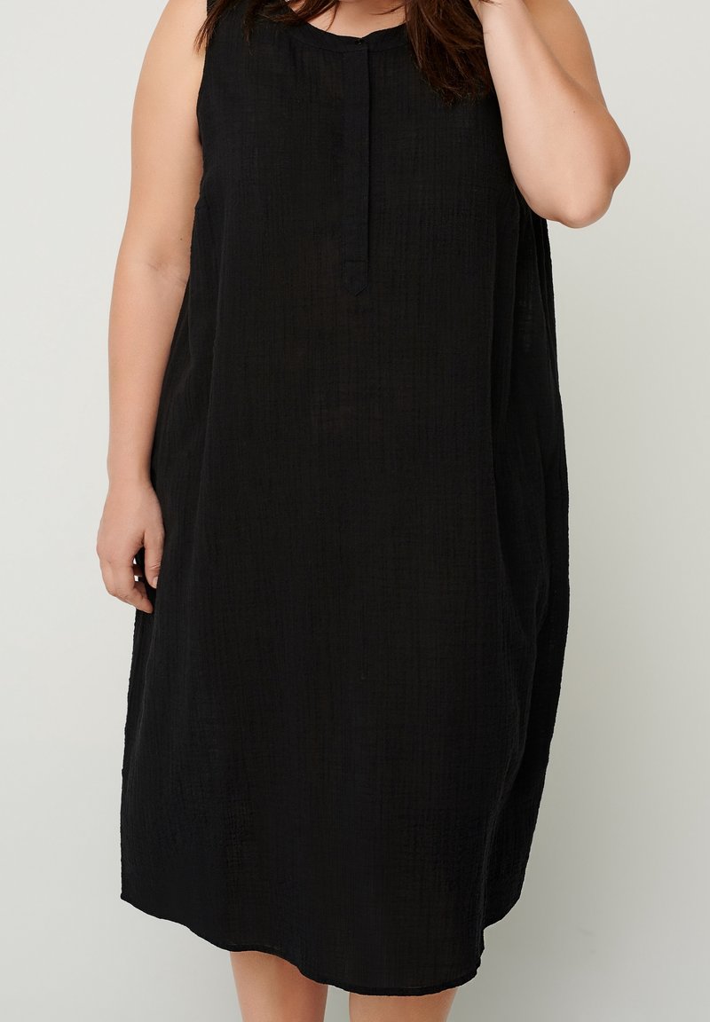 Femme portant une robe noire ample sans manches, longueur au genou, avec une texture subtile, debout devant un fond clair uni.