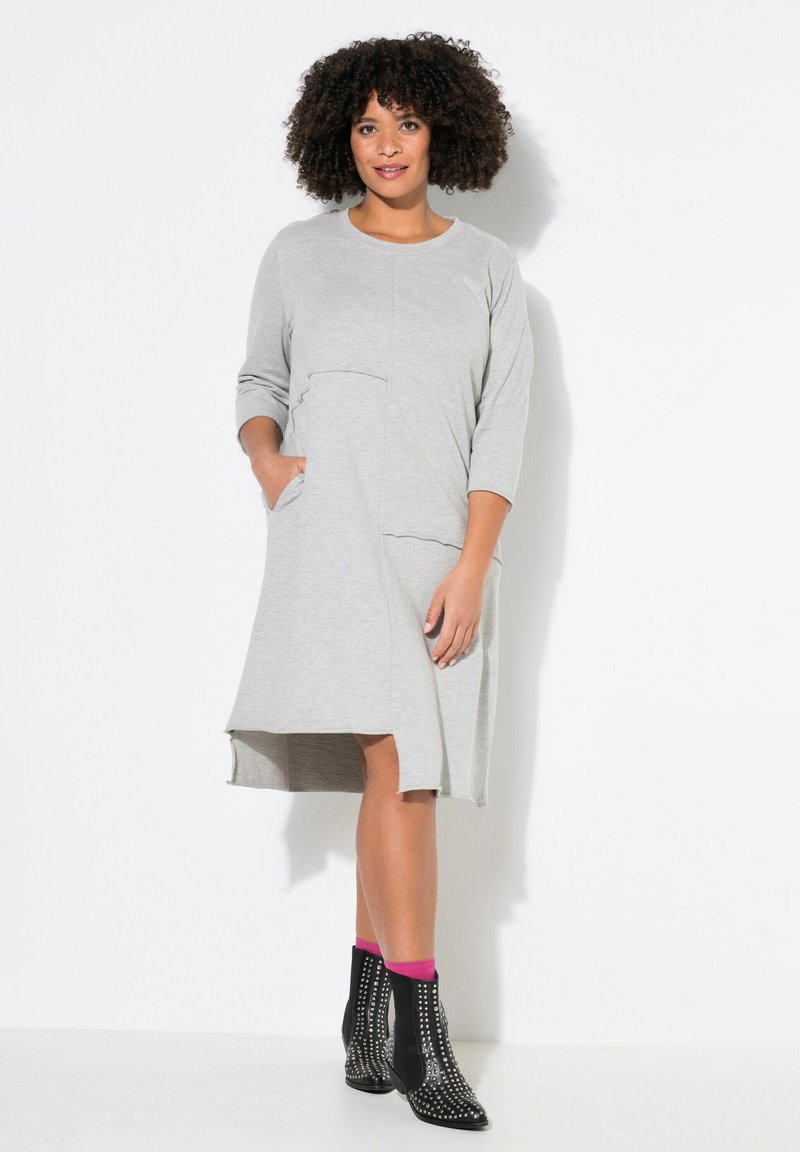 Robe grise longueur genoux avec des manches 3/4, un col rond et des poches latérales. Présente un ourlet légèrement asymétrique et un tissu texturé. Bottines noires avec clous.