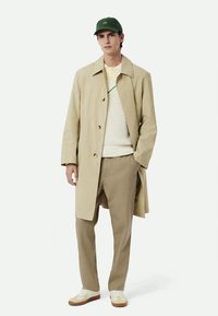 Palton bej, căciulă verde, pulover alb tricotat, pantaloni bej deschis și adidași albi cu talpă maro. Design simplu, fără modele.