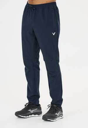 Homme portant un pantalon de sport bleu marine et des chaussures de course noires debout devant un fond blanc.