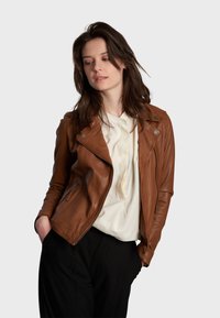 Giubbotto da motociclista in pelle marrone con tasche con zip, vestibilità aderente e dettagli in metallo argentato, abbinato a una blouse bianca e pantaloni neri.
