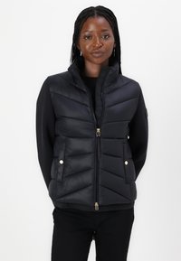 Vesta puffer neagră cu guler înalt, quilt diamantat, închidere prin fermoar frontal și buzunare cu capse de nuanță aurie. Purtați peste un top negru cu mânecă lungă.