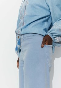 Chemise en denim bleu clair avec des détails brodés, accompagnée d'un pantalon bleu clair assorti. Les deux présentent une coupe décontractée et des accents à franges.