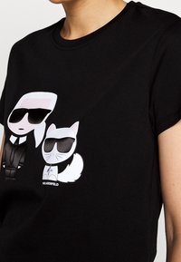 Camiseta negra con un diseño impreso de dos personajes: una persona con gafas de sol y un gato, ambos en estilo de dibujos animados.