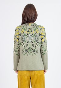 Pull en maille vert olive avec des motifs floraux jaunes et verts. Manches longues, col rond et texture lisse. Porté avec un pantalon en velours côtelé.