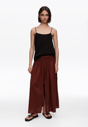 Mujer con top negro de tirantes finos, falda suelta de talle alto color granate con abertura frontal, y chanclas negras, de pie contra un fondo blanco.