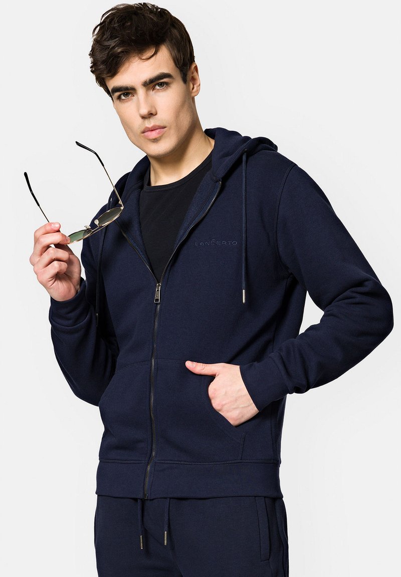 Granatowa bluza z kapturem na zamek, wykonana z miękkiego materiału, z kieszenią z przodu, kapturem ze sznurkiem i subtelnym haftem logo na klatce piersiowej.