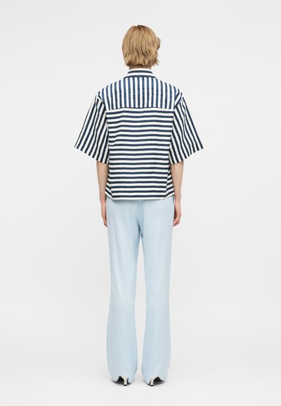 Άτομο που φοράει φαρδιά κοντομάνικη μπλούζα με μπλε και λευκές ρίγες και ανοιχτόχρωμο παντελόνι με φαρδιά μπατζάκια, στέκεται πλάτη σε απλό φόντο.