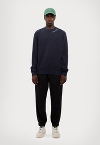 Granatowa bluza crewneck z logo w białym kolorze, czarne zwężane dresy i kremowe sneakersy, noszone z jasnobeżową czapką. Gładka faktura materiału.