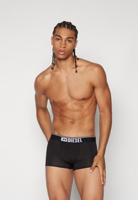 Black boxer briefs with a white Diesel logo on the waistband. Smooth texture and fitted design, model has braided hair and a toned physique.