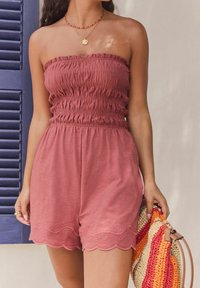Rosenfarbener trägerloser Jumpsuit mit gerafftem Oberteil, gewelltem Saum und bestickten Details, kombiniert mit einer mehrfarbigen geflochtenen Tasche.