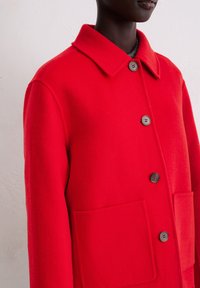 Rode wollen jas met een kraag, drie knopen en twee grote voorkantzakken. Zachte textuur met een getailleerde pasvorm.