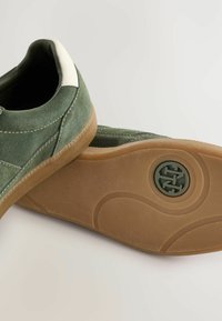 Baskets en suede vert avec un accent de talon blanc, semelle en caoutchouc au texture conçue, et détails de couture visibles.