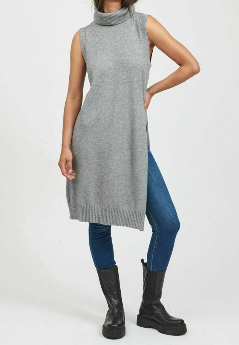 Grå ärmlös polotröja med ribbad krage och sidofickor, matchad med blå skinny jeans och svarta klackstövlar.