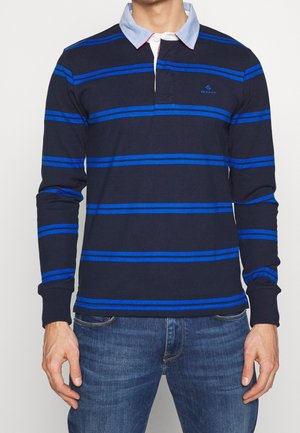 Mężczyzna ubrany w granatową koszulkę polo z długim rękawem, z jasnoniebieskimi poziomymi paskami i jasnoniebieskim kołnierzykiem, w połączeniu z niebieskimi dżinsami.