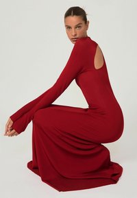Frau in einem langen roten gerippten Kleid mit langen Ärmeln und einem Rückenausschnitt, die sich vor einem schlichten hellen Hintergrund hockt.