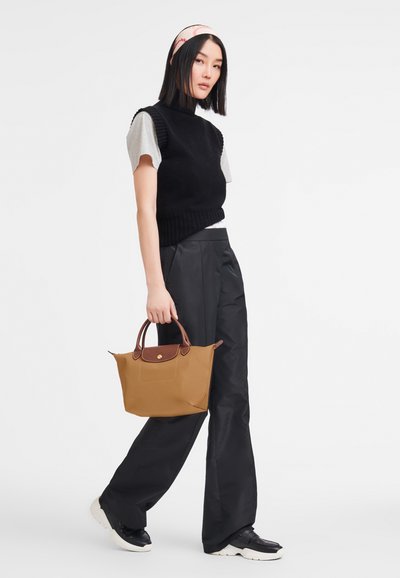 Tan tote bag with brown accents and a round handle. Model wears a black knit vest over a gray t-shirt and loose black pants, standing on a plain background.