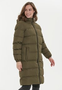 Abrigo acolchado de color verde oliva con capucha, cierre completo, puños elásticos y dos bolsillos laterales con cierre. Textura acolchada y diseño a la altura de la rodilla.